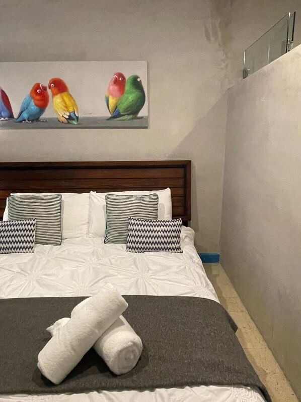 Una cama bien hecha con ropa blanca y almohadas a rayas, una manta gris y dos toallas blancas enrolladas. Sobre la cama, un cuadro de aves coloridas.