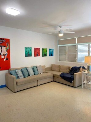 Un salón con un sofá seccional color beige, un ventilador de techo y cuatro cuadros con bombillas en la pared.