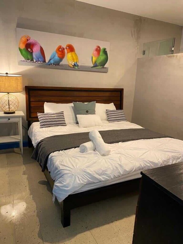 A hotel room with a large bed, a wooden headboard, a nightstand with a lamp, and a painting of colorful birds on the wall.