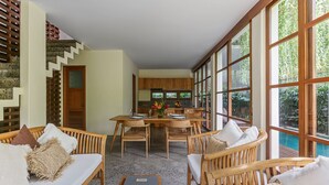 A spacious living area with a dining table, wooden furniture, and large windows overlooking a pool.