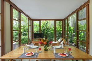 A dining area with a table set for two, a television, and a view of lush greenery through large windows. - Anandathu Villas (Canggu)