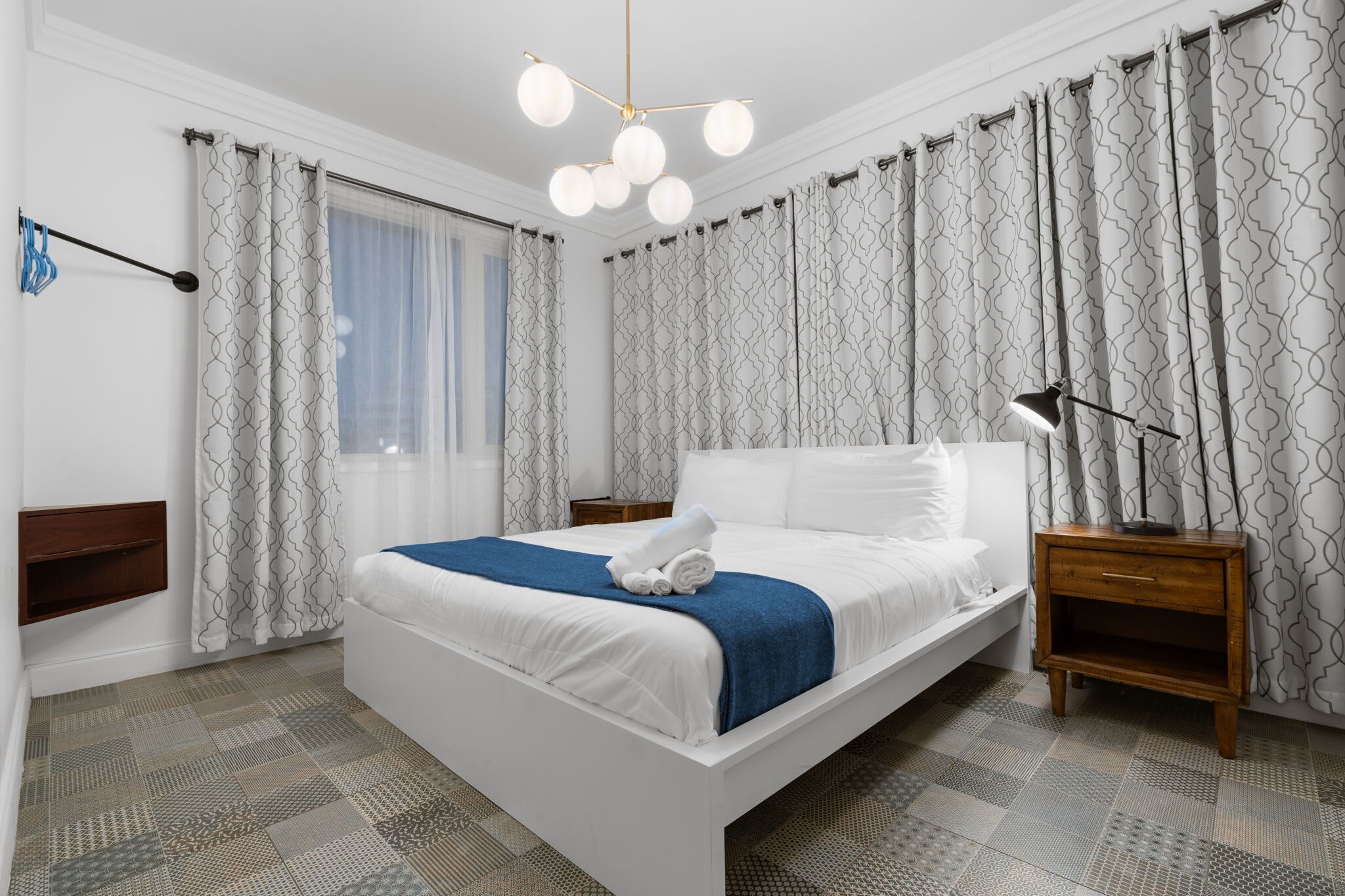 Une chambre moderne avec un grand lit, une table de chevet, une commode, un luminaire suspendu et des rideaux à motifs.