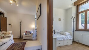 A room with a white sofa, a small white table, a patterned rug, a coat rack, and a window with a view of trees. - GuestHost - The window on the Raibl - Lovely APT with WiFi! x4 (Cave del Predil)