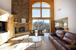 A spacious living room with a stone fireplace, a large window with a mountain view, a leather sofa, and a glass coffee table.