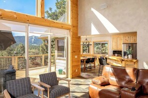 Amplia sala de estar con un ventanal con vistas a montañas, una terraza de madera y una cocina con gabinetes de madera.