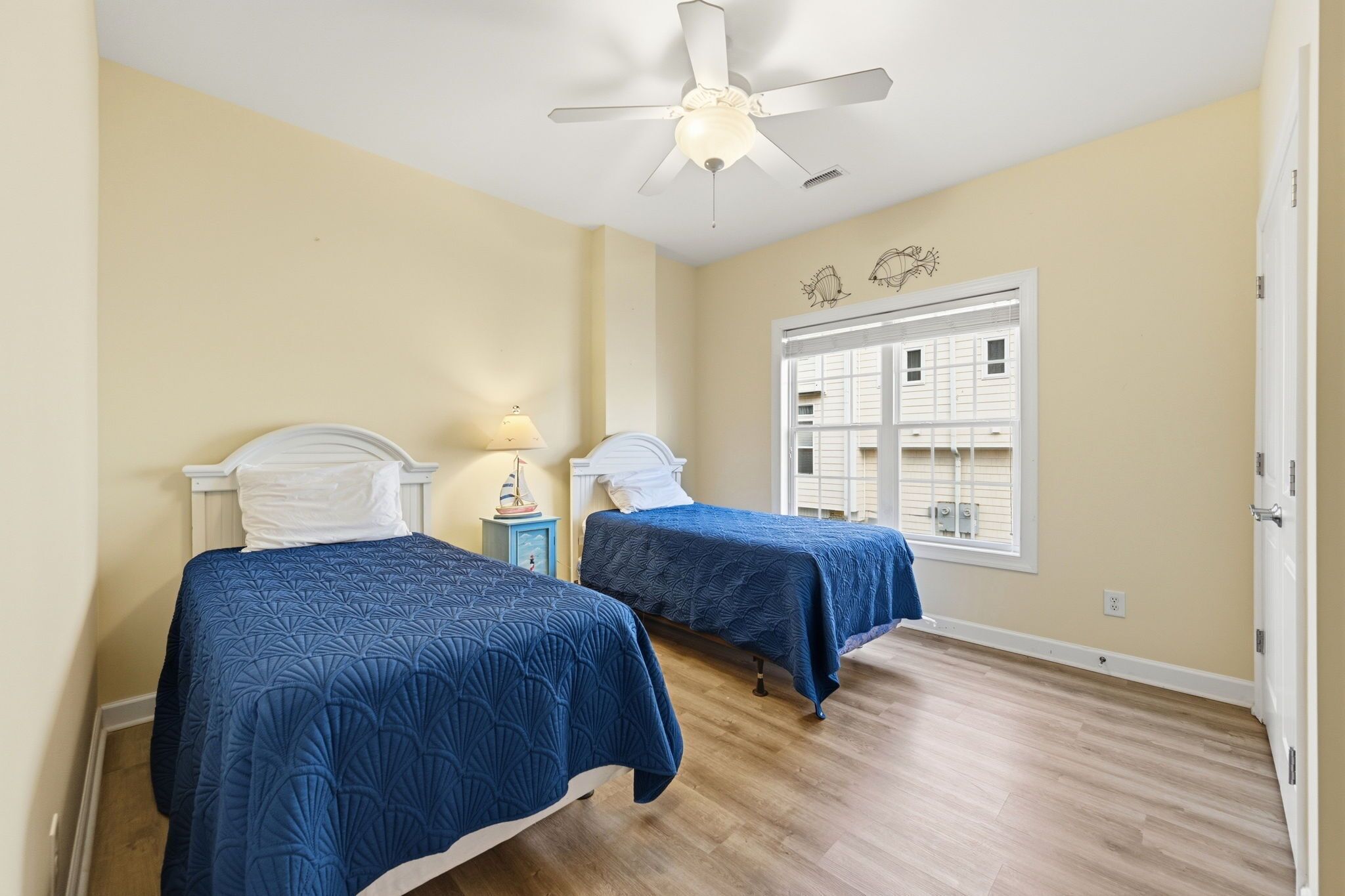 A room with two beds, a ceiling fan, a window with blinds, and a small nightstand with a lamp.