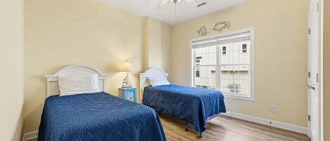 A room with two beds, a ceiling fan, a window with blinds, and a small nightstand with a lamp.