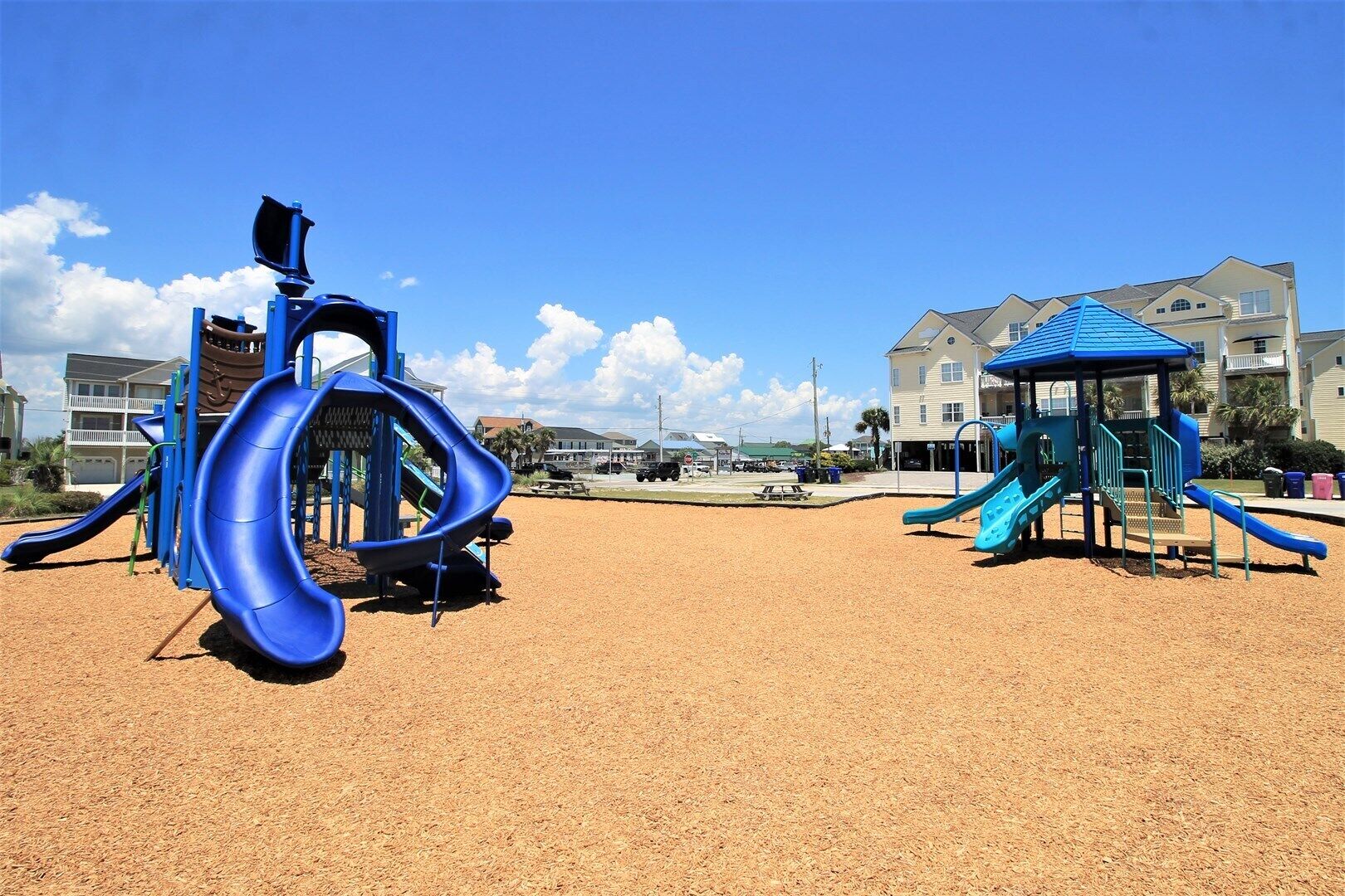 A playground with a blue slide and a blue climbing structure, set against a backdrop of residential buildings and a clear sky.