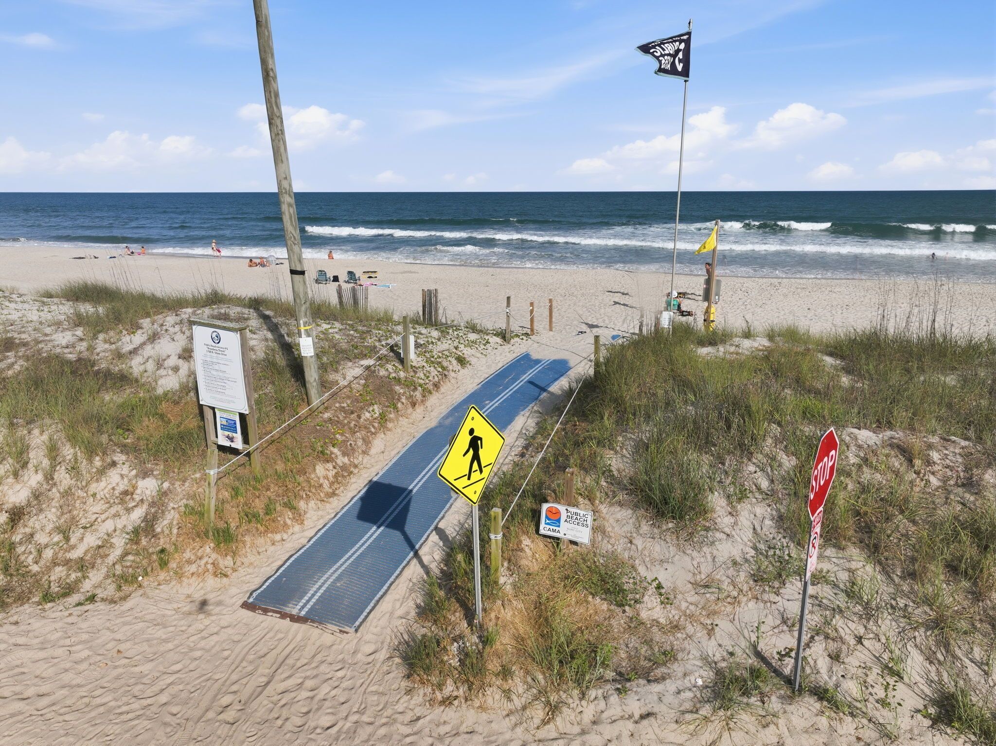 Un sentier menant à la plage, avec un panneau indiquant un passage piéton, un panneau de signalisation de stop et un drapeau.