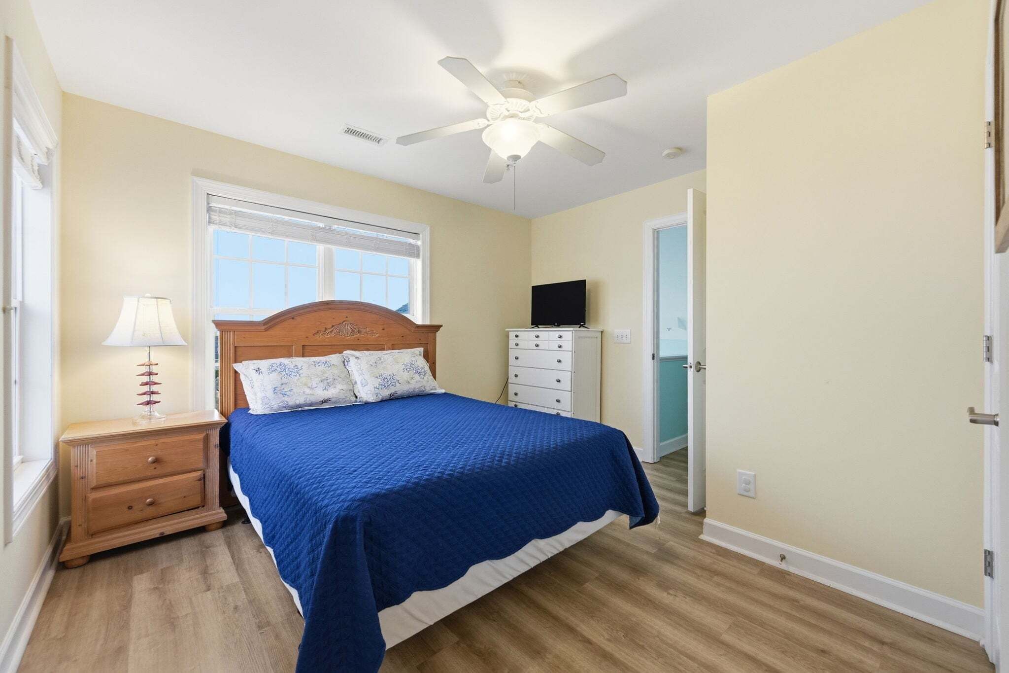 A bedroom with a bed, a nightstand, a lamp, a ceiling fan, a TV, and a window.