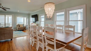 A dining area with a wooden table and white chairs, a chandelier, and large windows with blinds. - Sandy Paradise: An oceanfront, 6-bedroom retreat in Surf City, perfect for 20 guests! (Surf City)