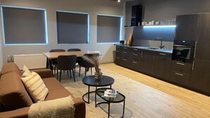 A modern living room with a brown sofa, a dining table, and a kitchen area with dark cabinets and a built-in oven.