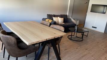 A modern living room with a wooden dining table, cushioned seating, a small side table, and a flat-screen TV mounted on the wall.