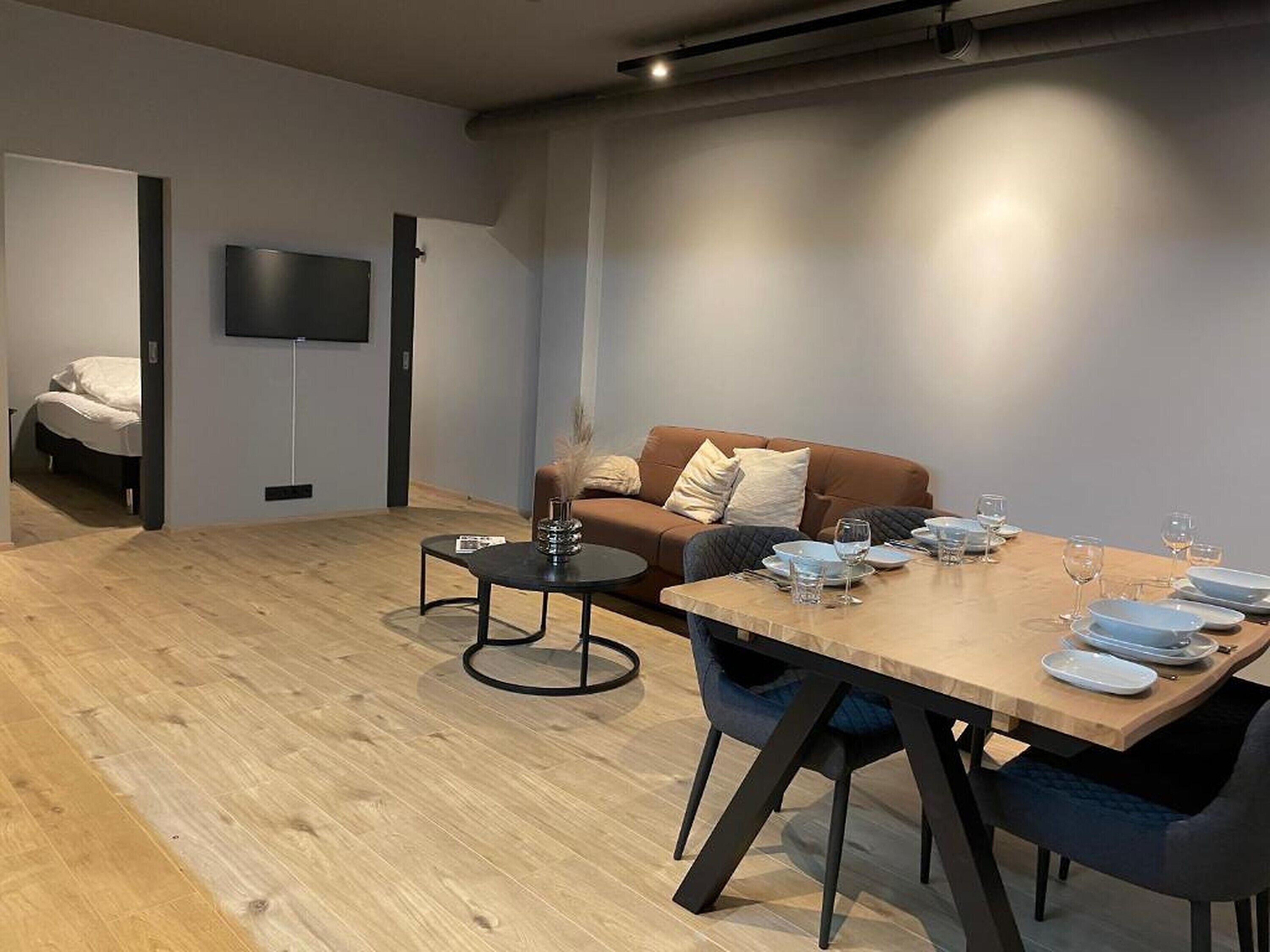 Un soggiorno moderno con un tavolo da pranzo in legno apparecchiato per quattro persone, un divano marrone e un televisore a schermo piatto montato a parete.