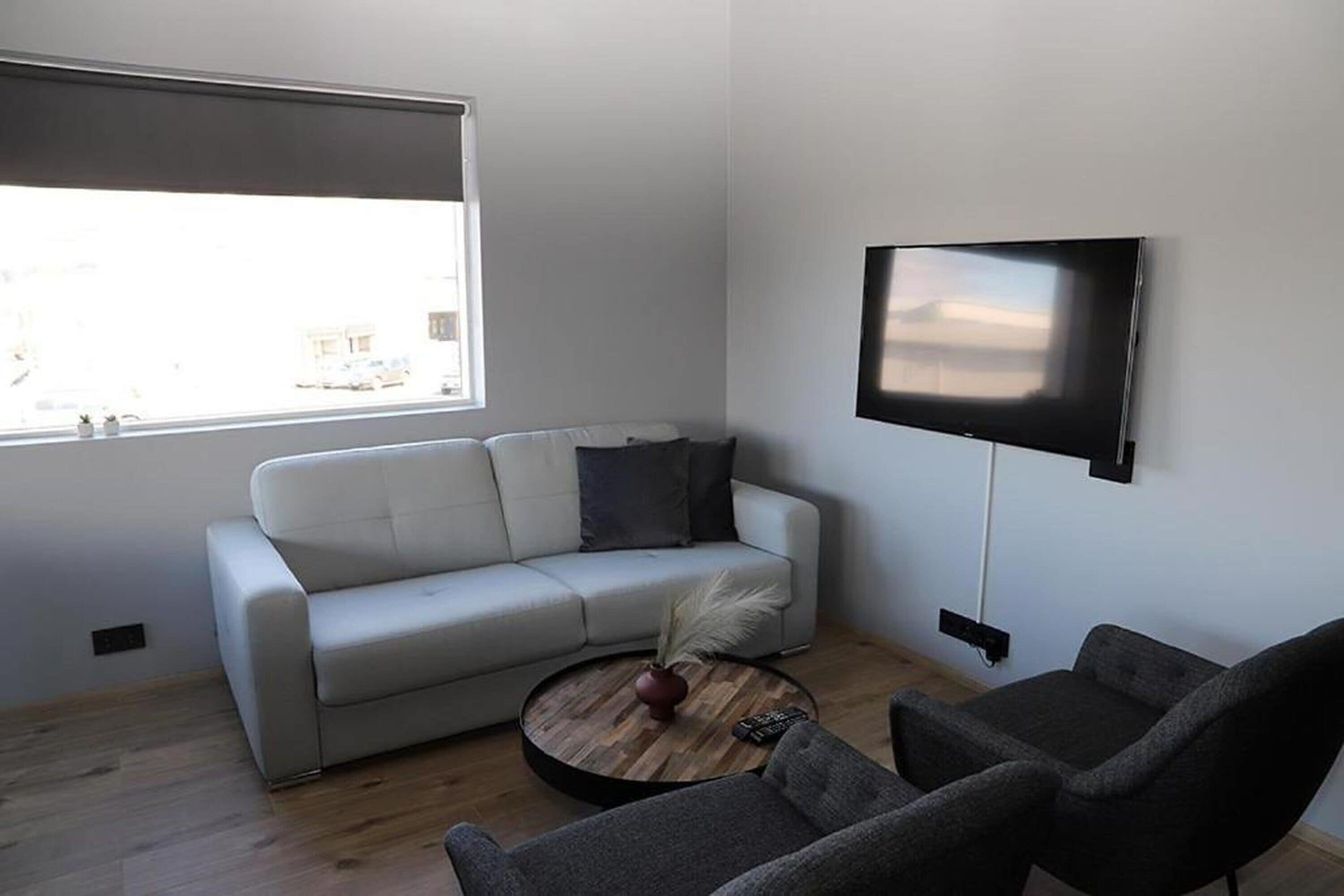 Ein modernes Wohnzimmer mit einem weißen Sofa, einem runden Couchtisch mit einer Vase und Federn, einem grauen Sessel und einem flachbildfernseher an der Wand.