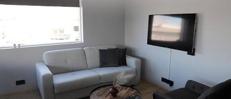 A modern living room with a white sofa, a round coffee table with a vase and feathers, a grey armchair, and a flat-screen TV mounted on the wall.