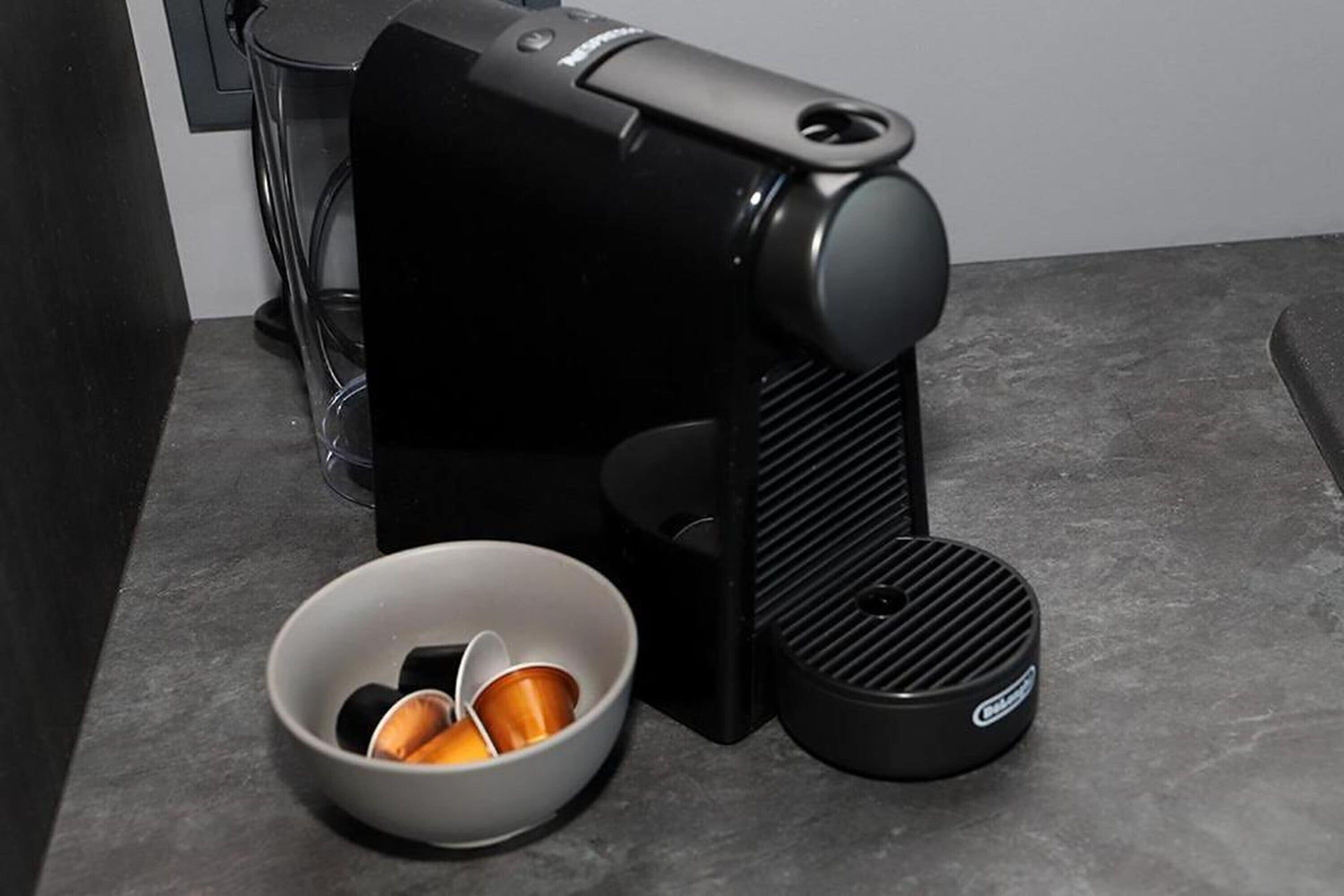 A black coffee machine with a cup holder and a bowl of coffee pods on a countertop.