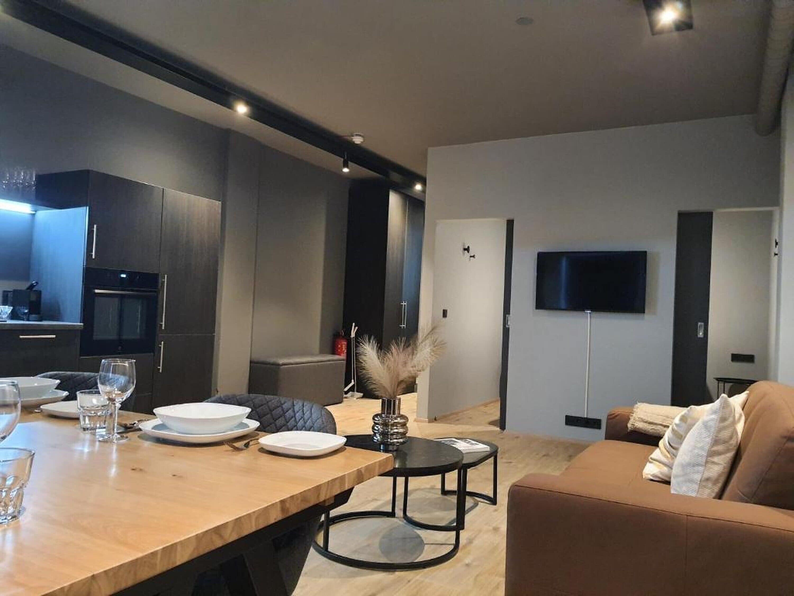 Uma sala de estar moderna com uma mesa de jantar de madeira posta para dois, um sofá e uma TV de tela plana na parede.