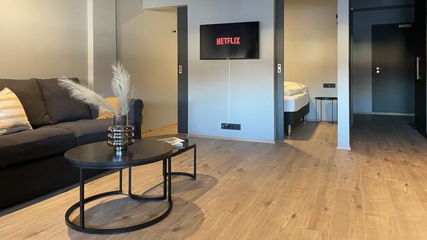 A modern living room with a sofa, a coffee table, a TV mounted on the wall, and a door leading to a bedroom.