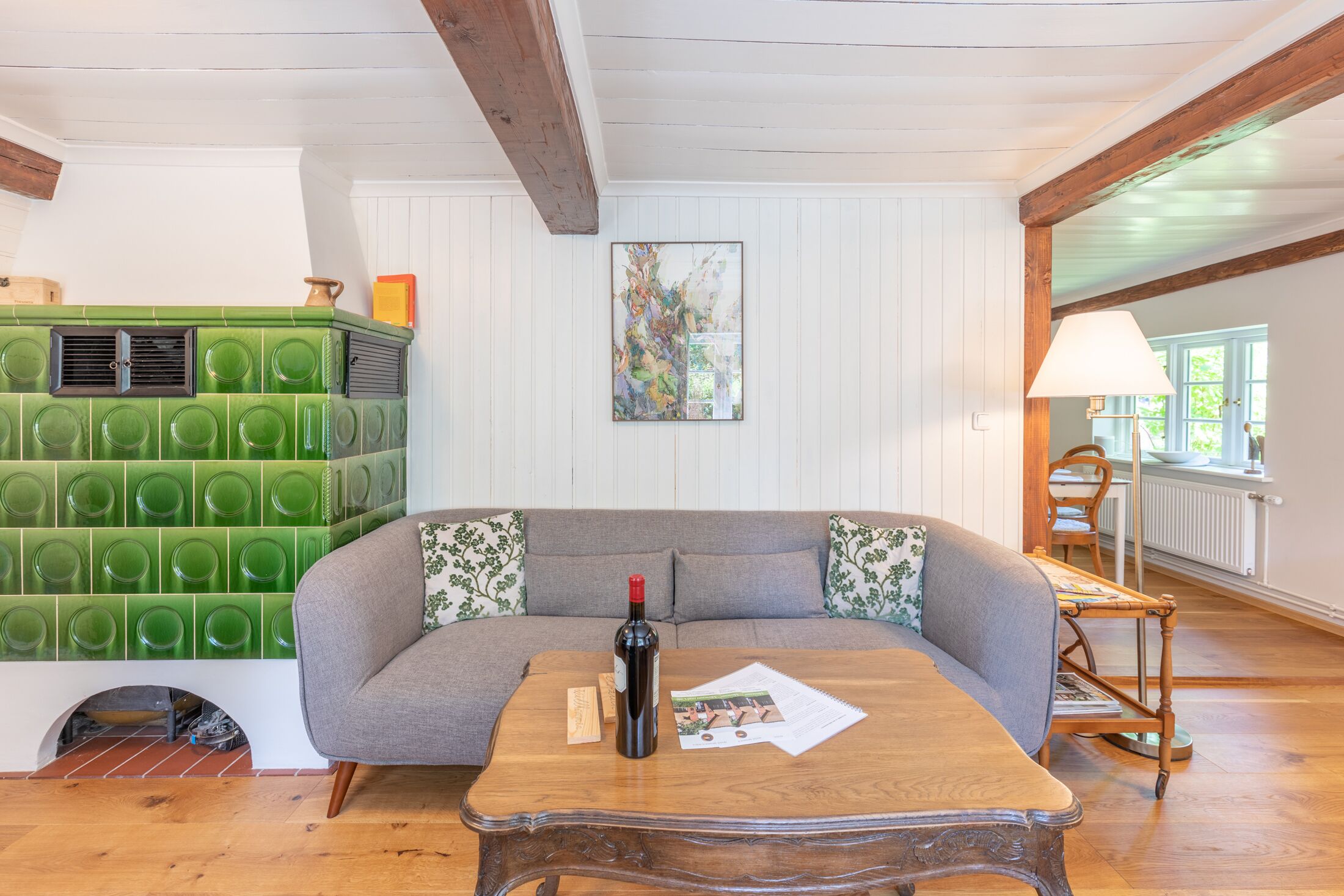 A cozy living room with a wooden coffee table, a sofa with floral cushions, a green tiled fireplace, and a painting on the wall.