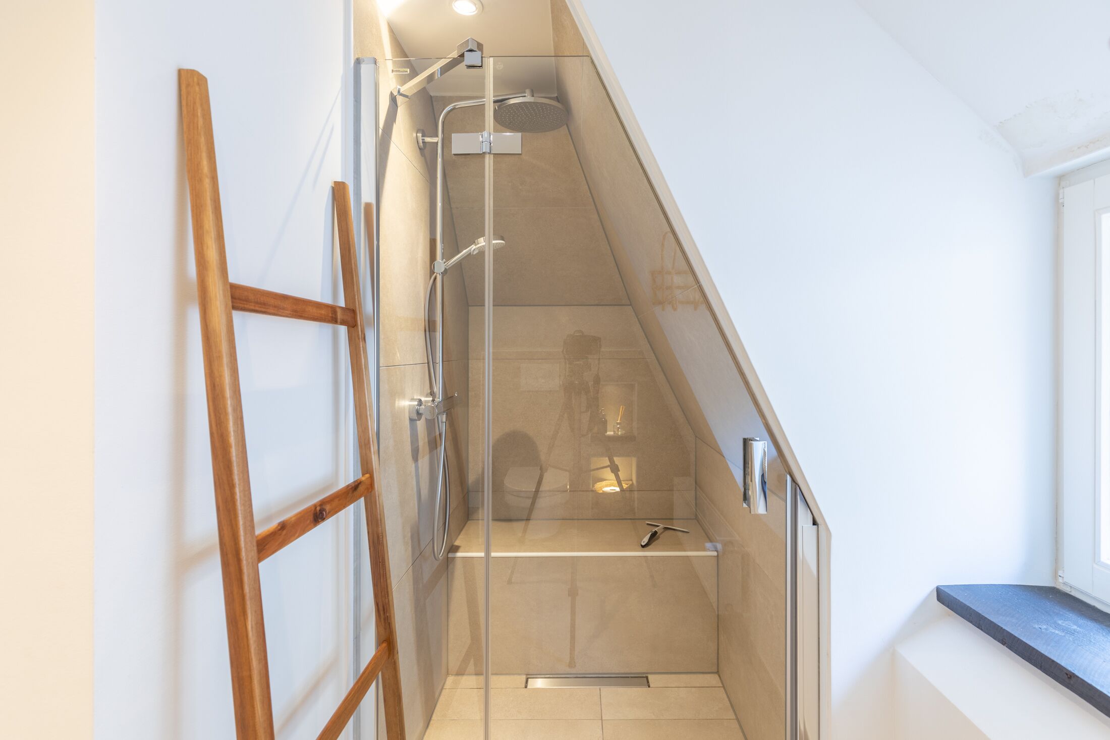 Ein modernes Badezimmer mit Duschkabine aus Glas, einer Holzleiter und einem kleinen Fenster.