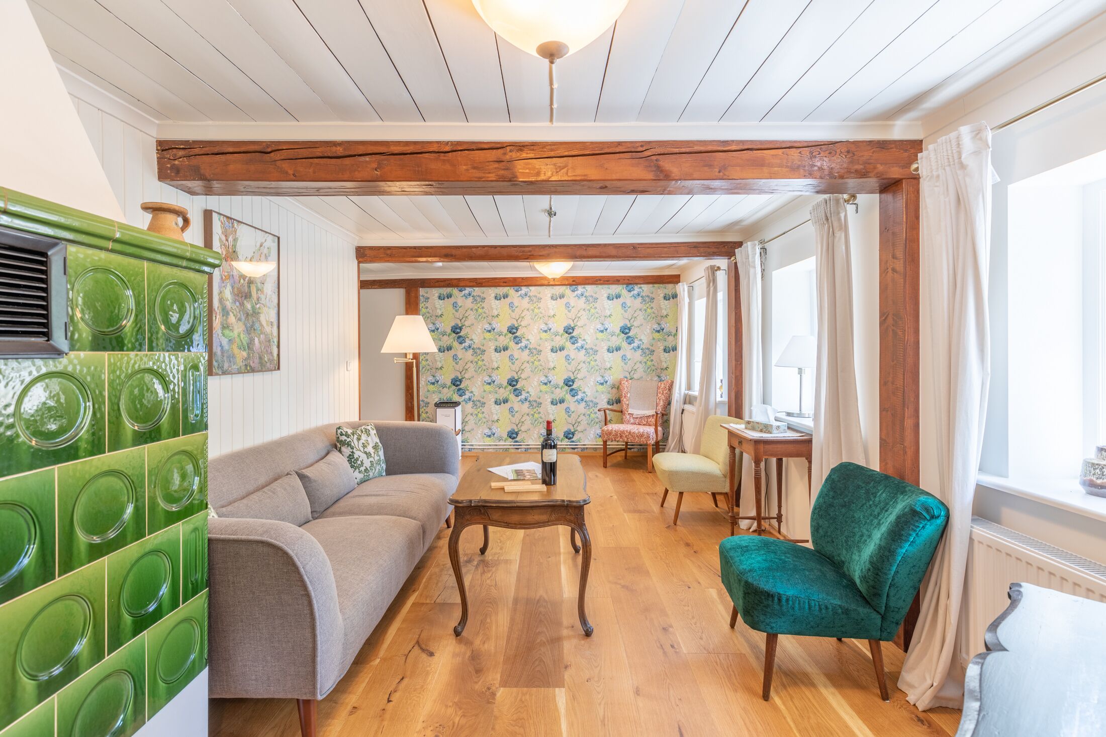 Een gezellige woonkamer met een houten plafond, een groene betegelde muur, een bank, een groene fauteuil en een tafel met een fles en een boek.