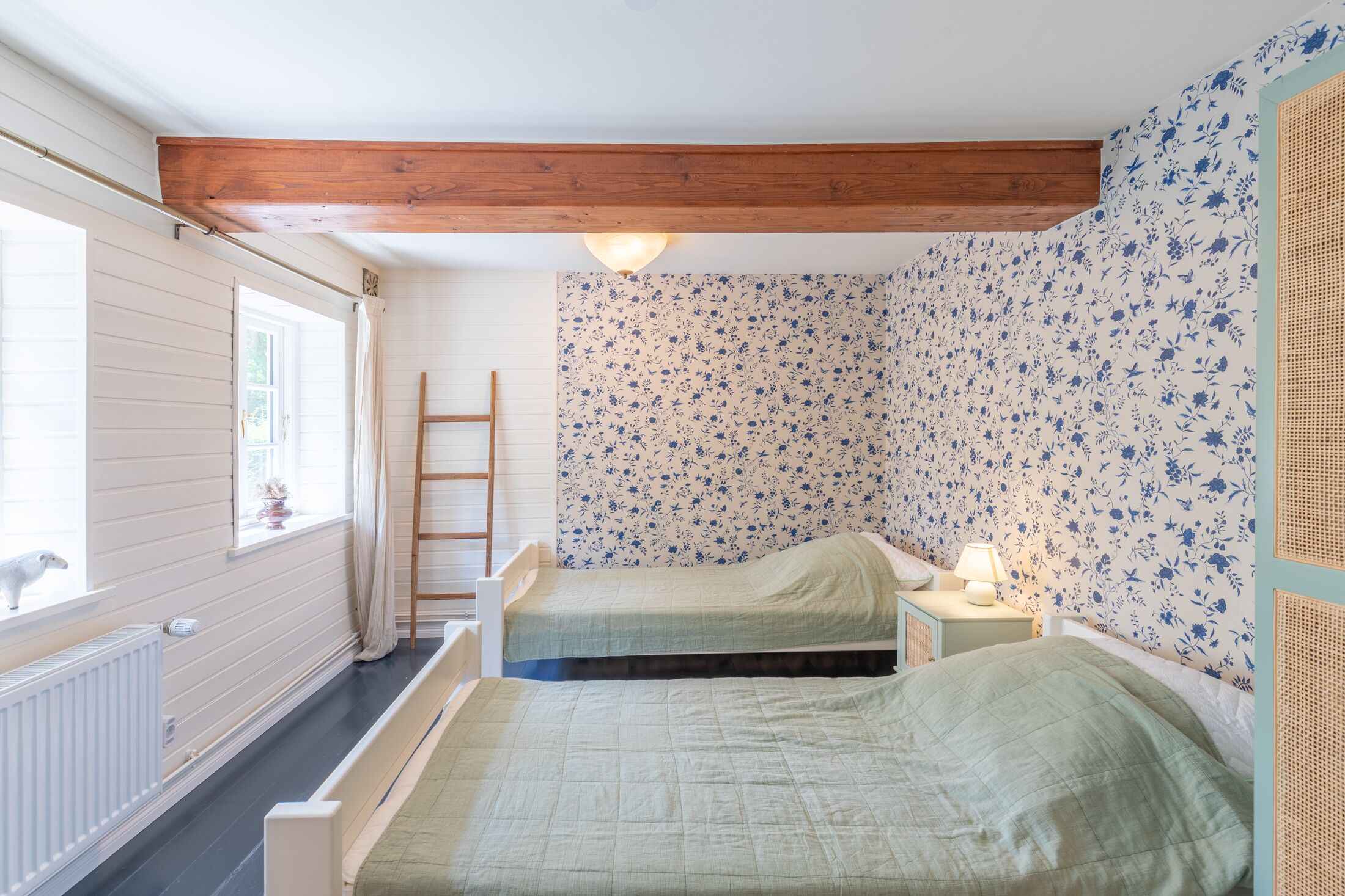 A bedroom with two beds, a wooden ceiling, a window with curtains, and a wall with floral wallpaper.