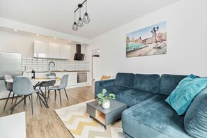 A modern living room with a blue sofa, a dining table with chairs, a kitchen in the background, and a cityscape painting on the wall.