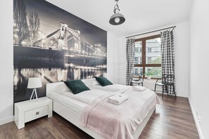 A bedroom with a large bed, a nightstand, a lamp, a chair, a window with curtains, and a mural of a bridge. - Stylish Apartment with Parking | Close to Market Square (Wrocław)