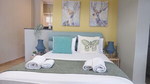 A neatly made bed with a green blanket, two pillows, and two rolled towels. There are two wall-mounted lamps and a potted plant on a side table. - Check Point - Museum (Athens)