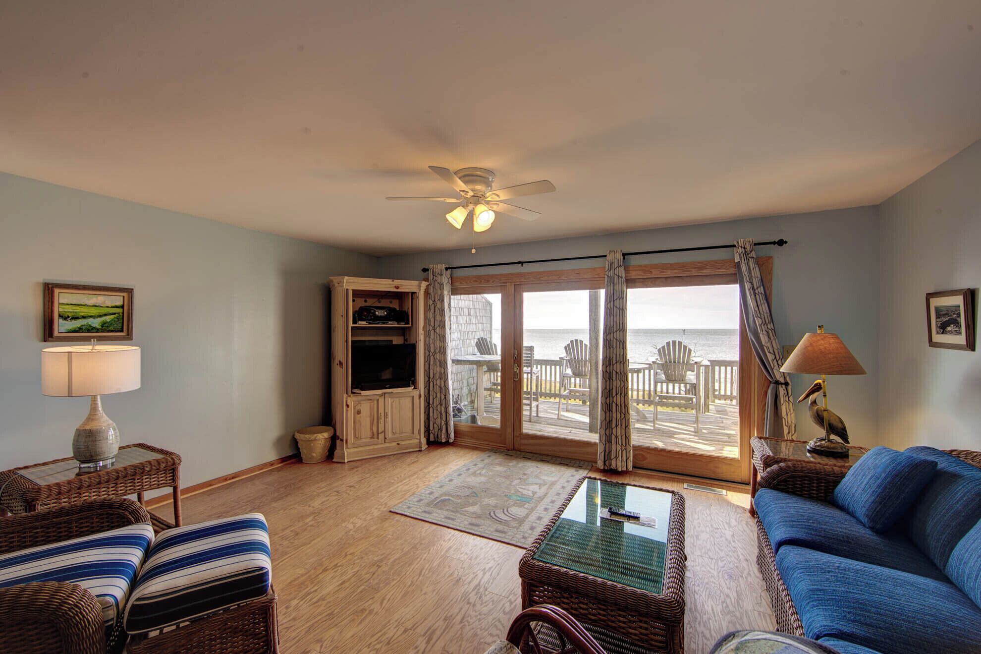 A living room with a large sliding glass door leading to a deck with chairs and a view of the ocean.