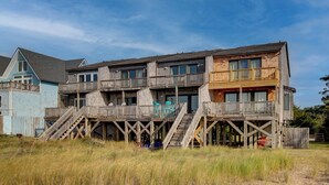 A multi-story wooden house with a deck and stairs, surrounded by grass.