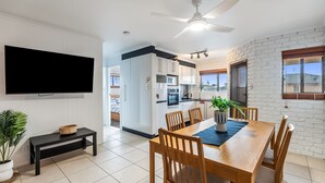 Une cuisine moderne avec des meubles blancs, un coin repas comprenant une table et des chaises en bois, un ventilateur de plafond et un téléviseur fixé au mur.