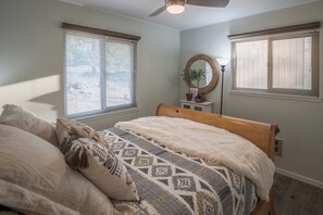 Un dormitorio con una cama, dos ventanas con persianas, un ventilador de techo, una mesita de noche con lámpara y un espejo montado en la pared.