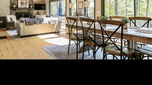 Amplia sala de estar con techo alto, chimenea, una gran mesa de comedor con sillas y ventanas amplias que ofrecen vistas a árboles.