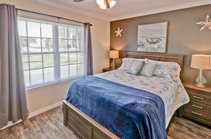 A bedroom with a large bed, a window with blinds, a starfish decoration, a painting, and a nightstand with a lamp.