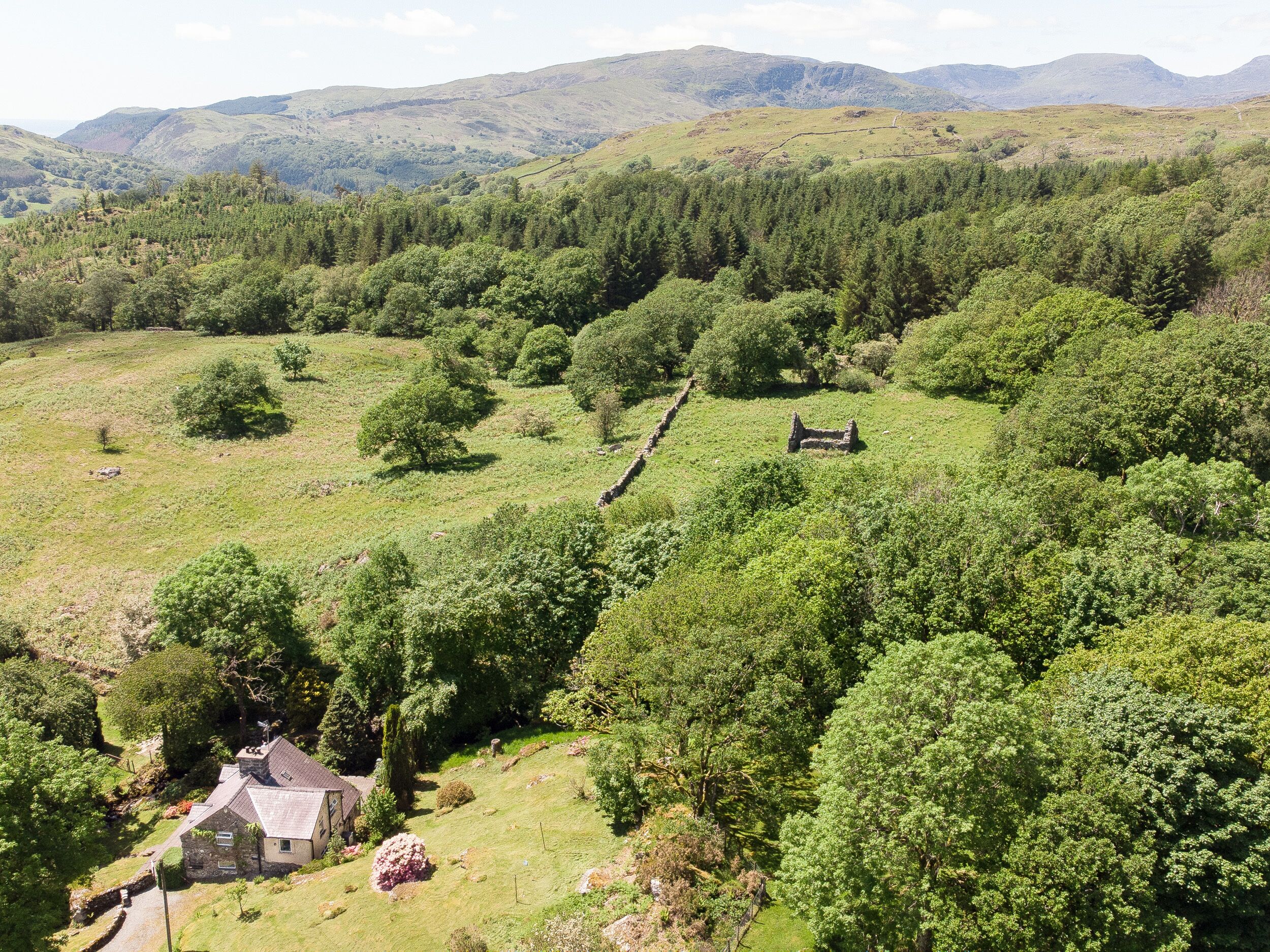 Une maison en pierre, dotée d’un toit à pente, entourée de champs verdoyants et de forêts denses, avec des montagnes en toile de fond.