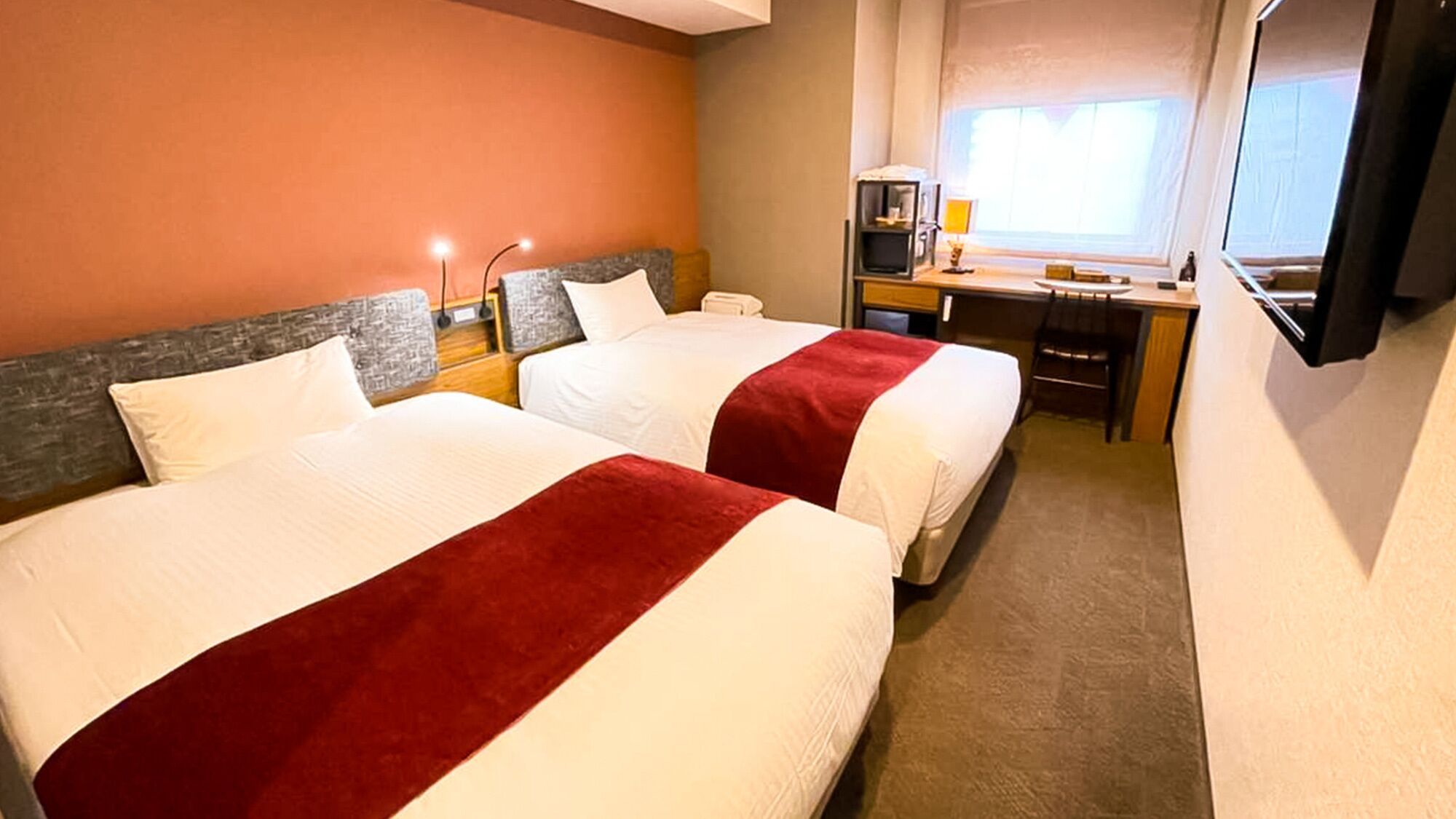 A hotel room with two beds, a desk, a chair, a TV, and a window.