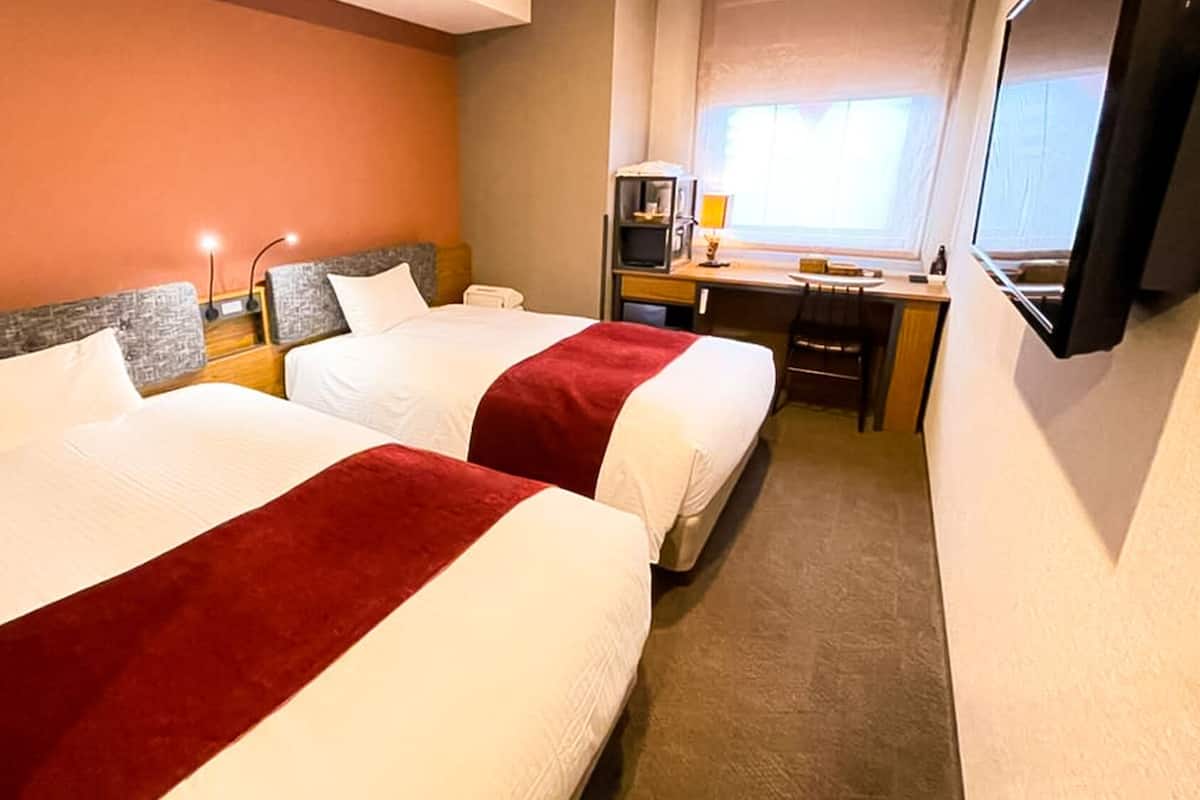 A hotel room with two beds, a desk, a chair, a TV, and a window.