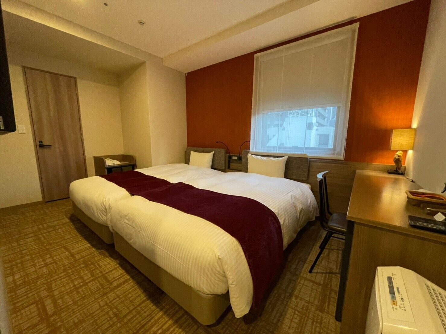 Quarto de hotel com uma cama grande, uma escrivaninha, uma cadeira, uma janela e uma porta.