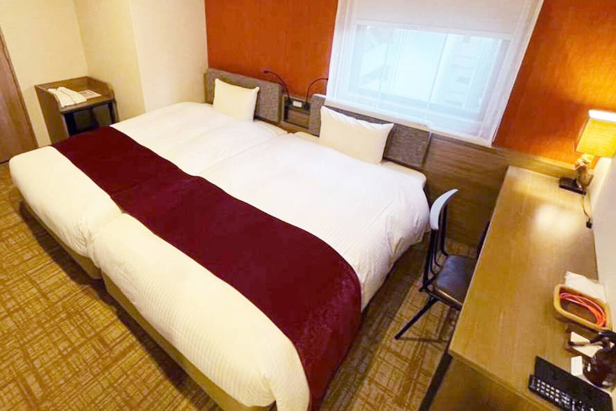 A hotel room with a bed, a desk, a chair, a nightstand, and a window.
