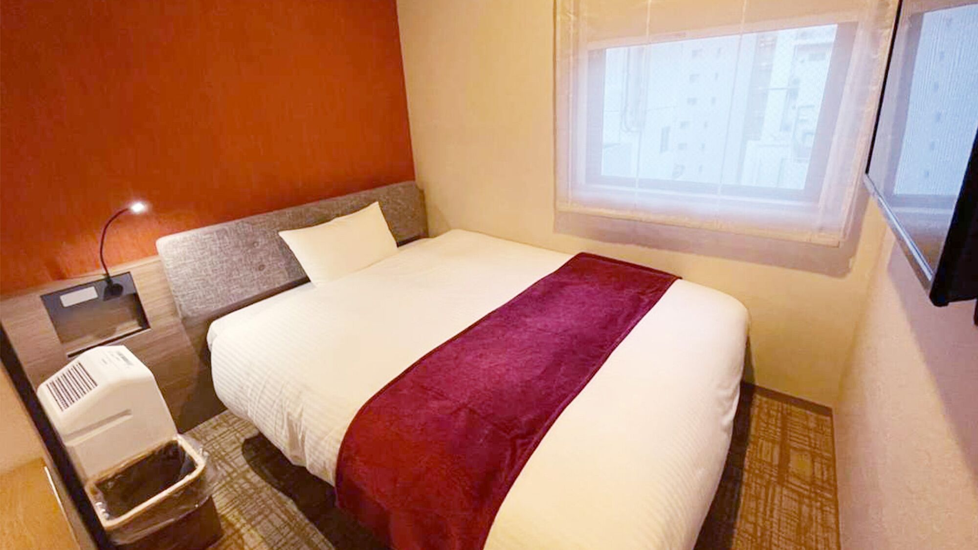 A hotel room with a bed, a window, a wall-mounted heater, and a trash bin.