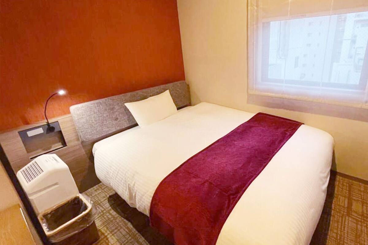 A hotel room with a bed, a window, a wall-mounted heater, and a trash bin.