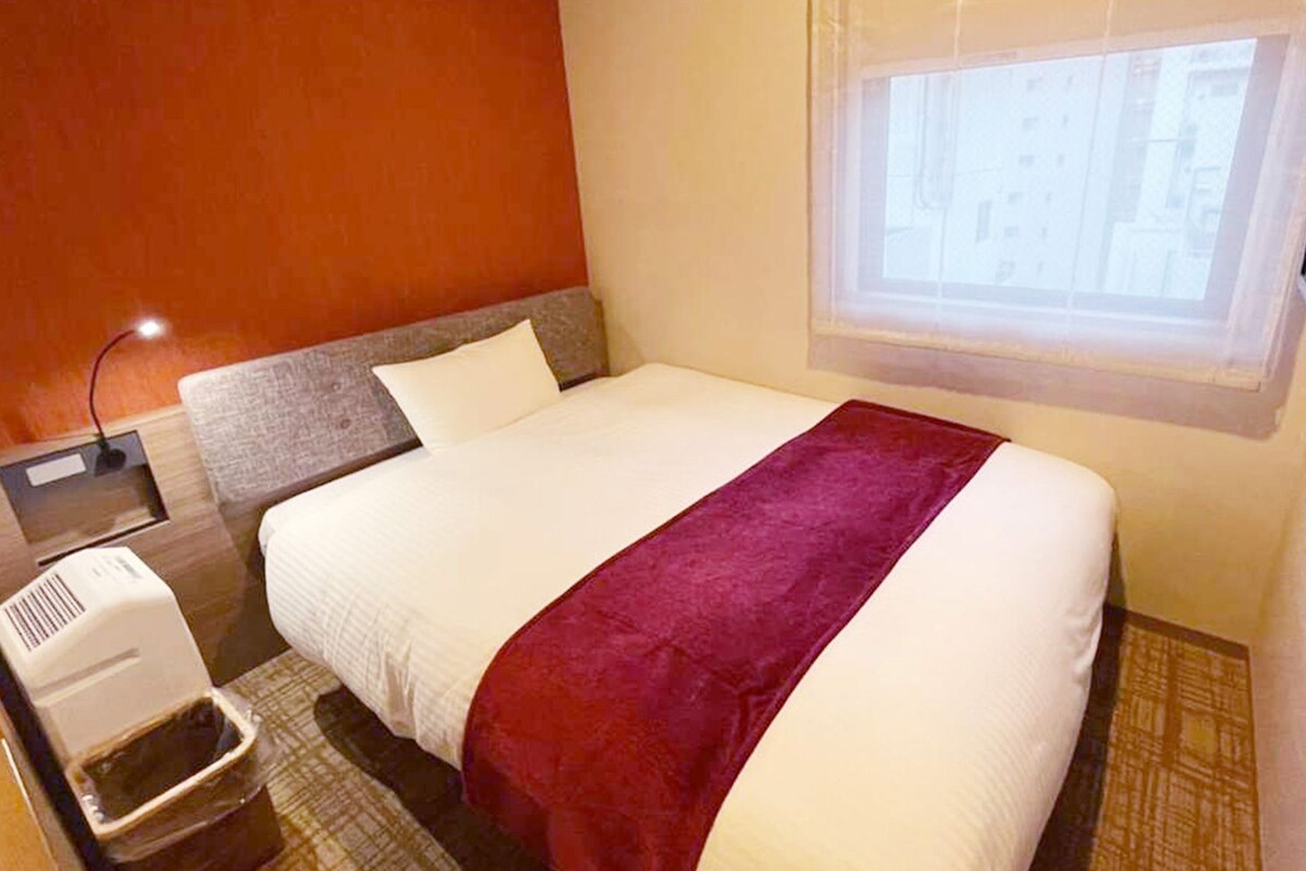 A hotel room with a bed, a window, a wall-mounted heater, and a small table with a heater.