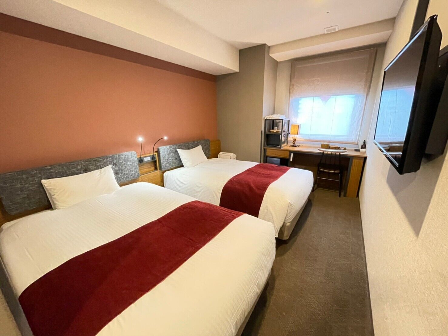 Quarto de hotel com duas camas, uma escrivaninha, uma televisão e uma janela.