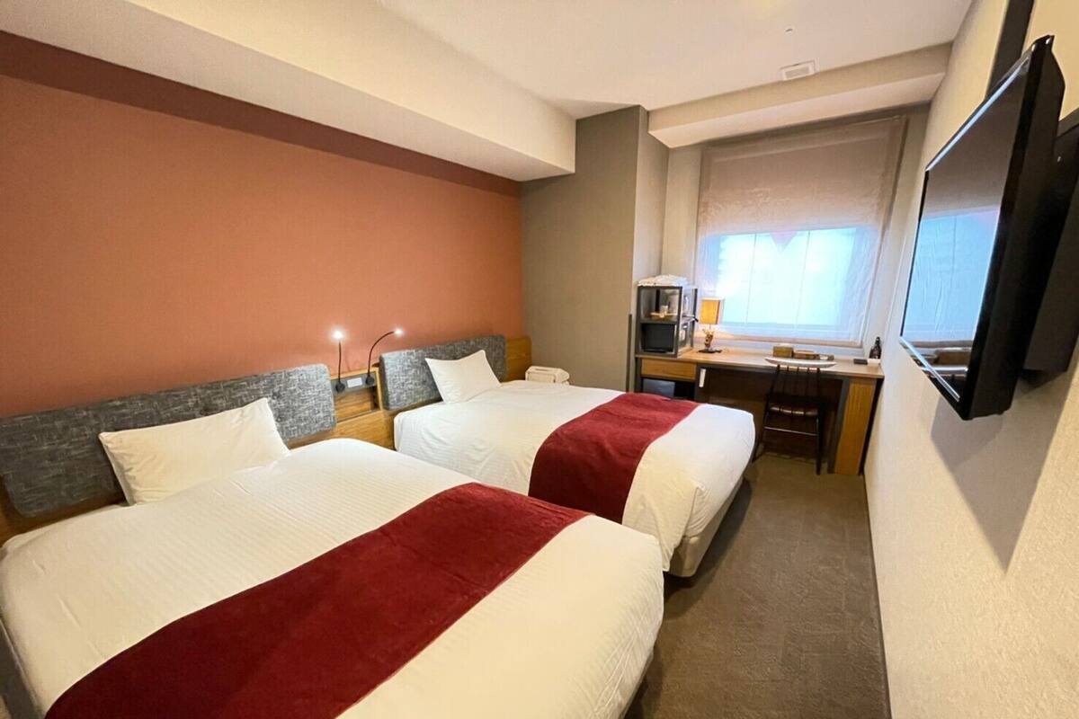 A hotel room with two beds, a desk, a TV, and a window.