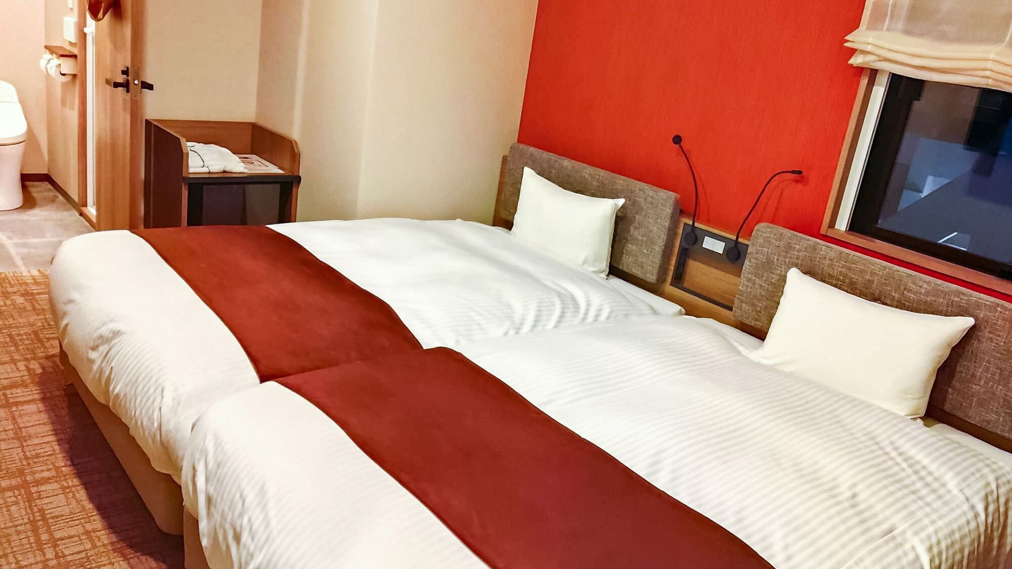 A hotel room with a bed, a nightstand, a window, and a red wall.