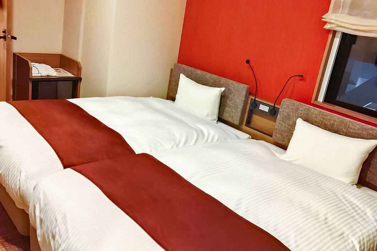 A hotel room with a bed, a nightstand, a window, and a red wall.