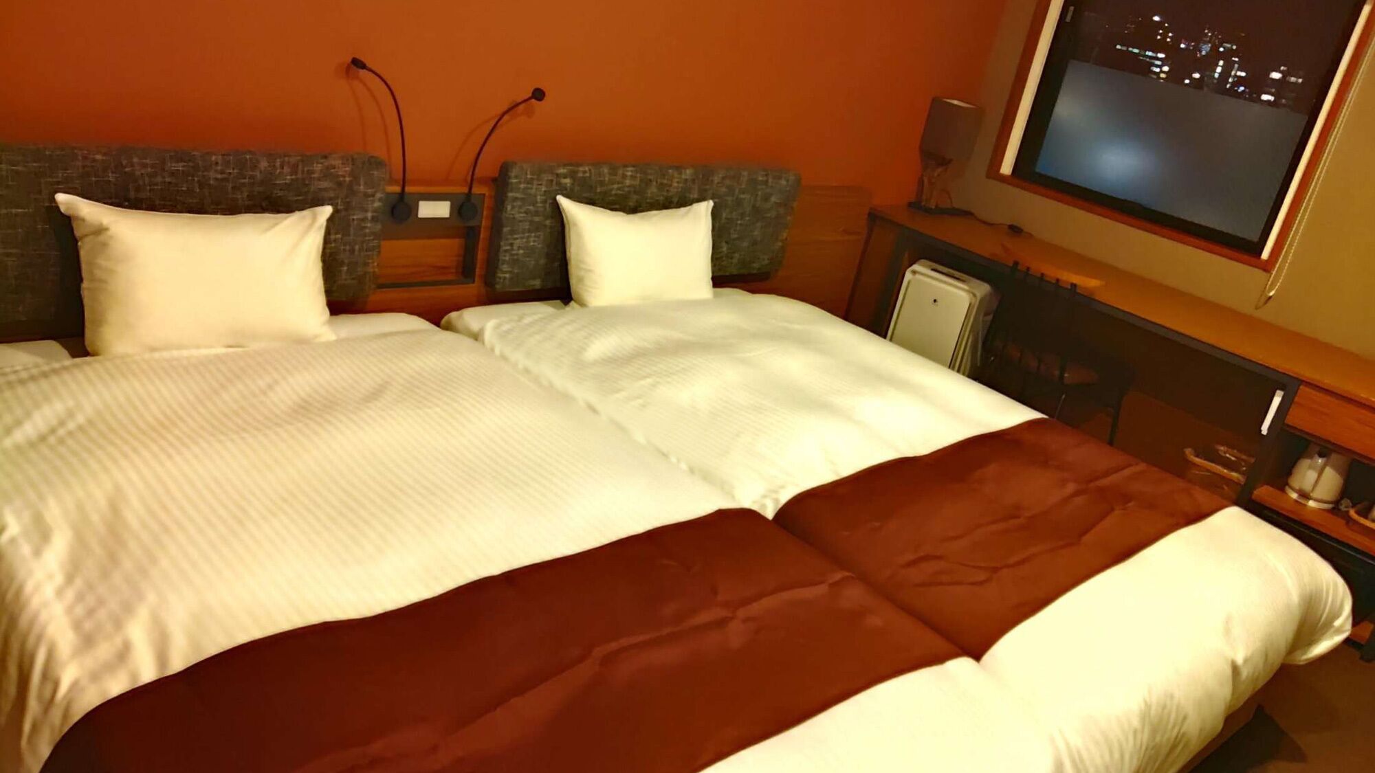 Uma cama de casal com roupa de cama branca e castanha, uma televisão na parede e vista para a cidade.