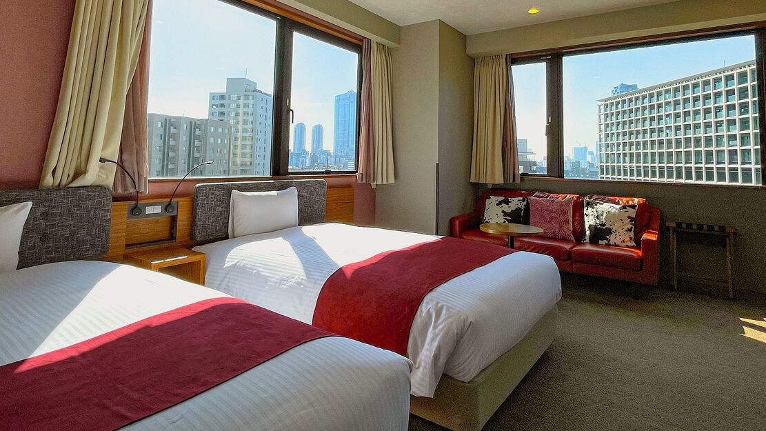 A hotel room with two beds, a sofa, a small table, and large windows with a city view.